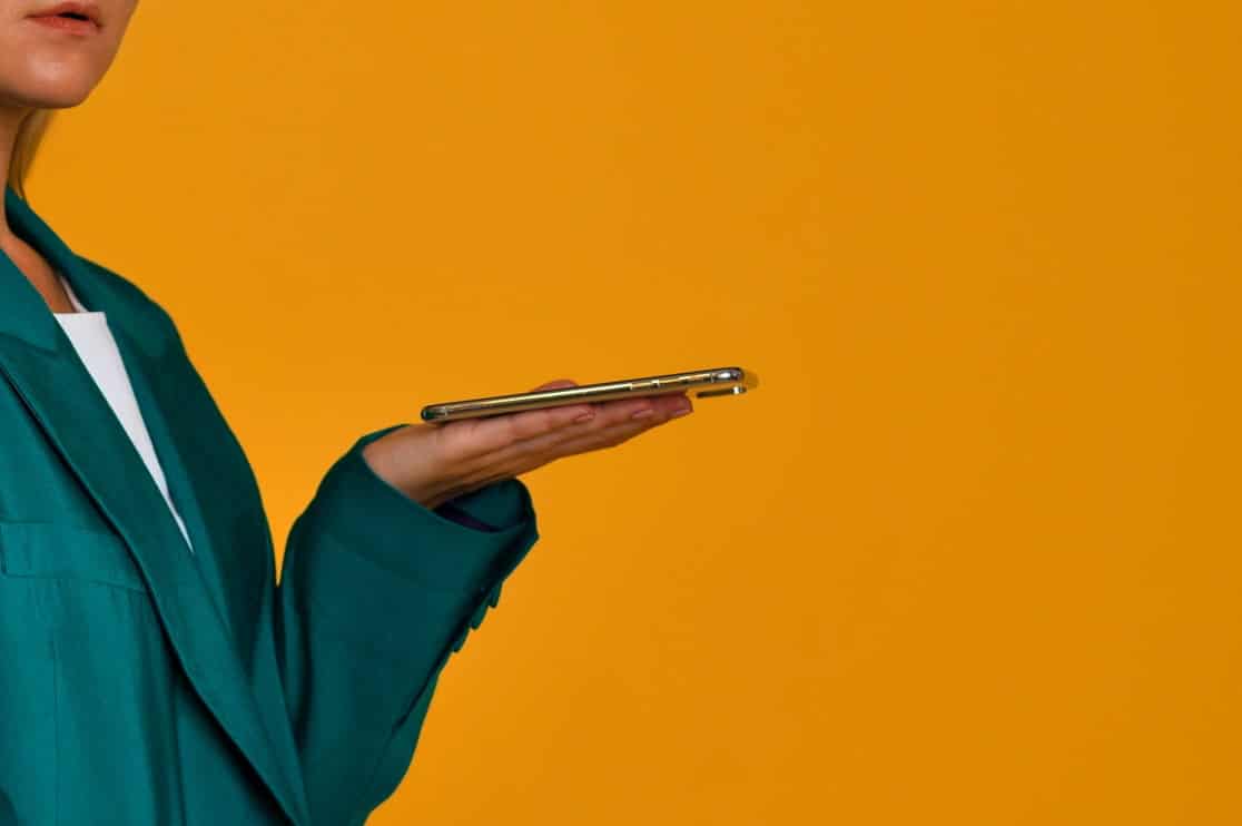 A person wearing a teal blazer holds a smartphone horizontally on their open palm against a solid yellow background. Only the lower half of their face and upper body are visible.