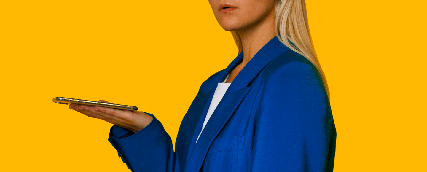 A person with long blonde hair wearing a blue blazer holds a smartphone on their open palm, speaking to it, against a solid yellow background.