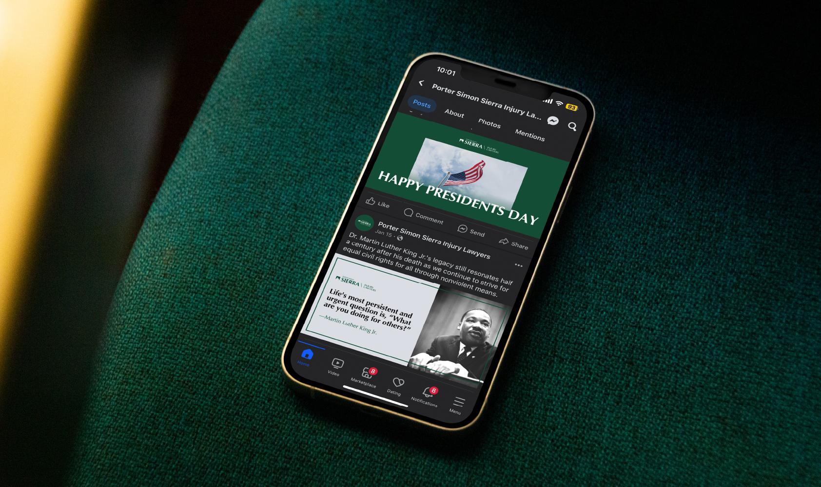 A smartphone on a green fabric surface displays a Facebook page, featuring a post with the text HAPPY PRESIDENTS DAY and an image of the U.S. flag, along with a post showing a quote and photo of Martin Luther King Jr.