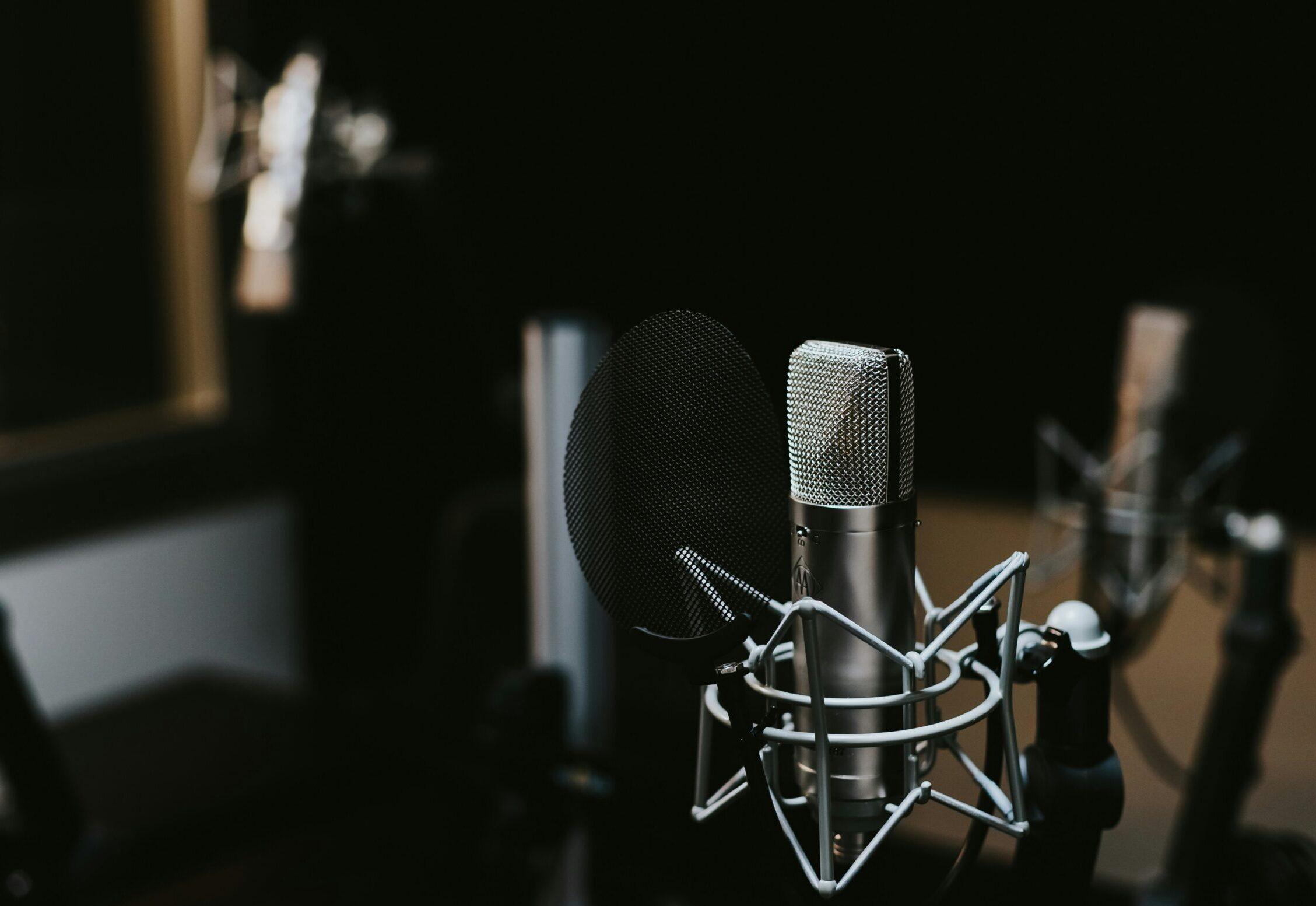 A professional microphone with a pop filter is set up in a recording studio, surrounded by other audio equipment, against a dark background.