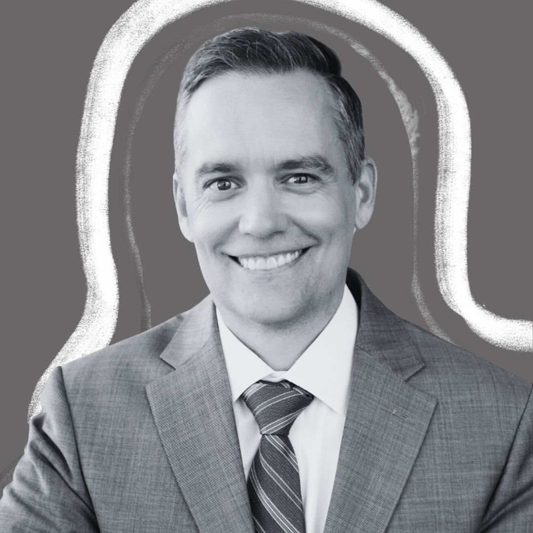 A man in a suit and tie smiles at the camera. The image is in grayscale, and there is a white, curved outline around his head and shoulders, set against a plain gray background.