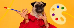 A pug wearing a red shirt is held upright by human hands. One hand holds a paintbrush, and a colorful artists palette is shown on a bright yellow background, highlighting the influence of color psychology in art.