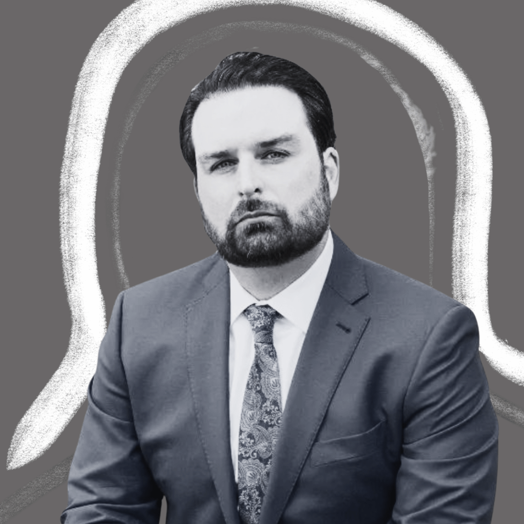A man with dark hair and a beard, wearing a suit and patterned tie, sits against a gray background with a white, abstract outline around his head and shoulders.